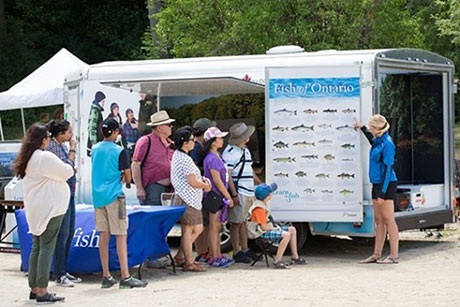 Image of an instructor outdoors pointing to a Fish of Ontario poster on a trailer in front of an audience of participants 