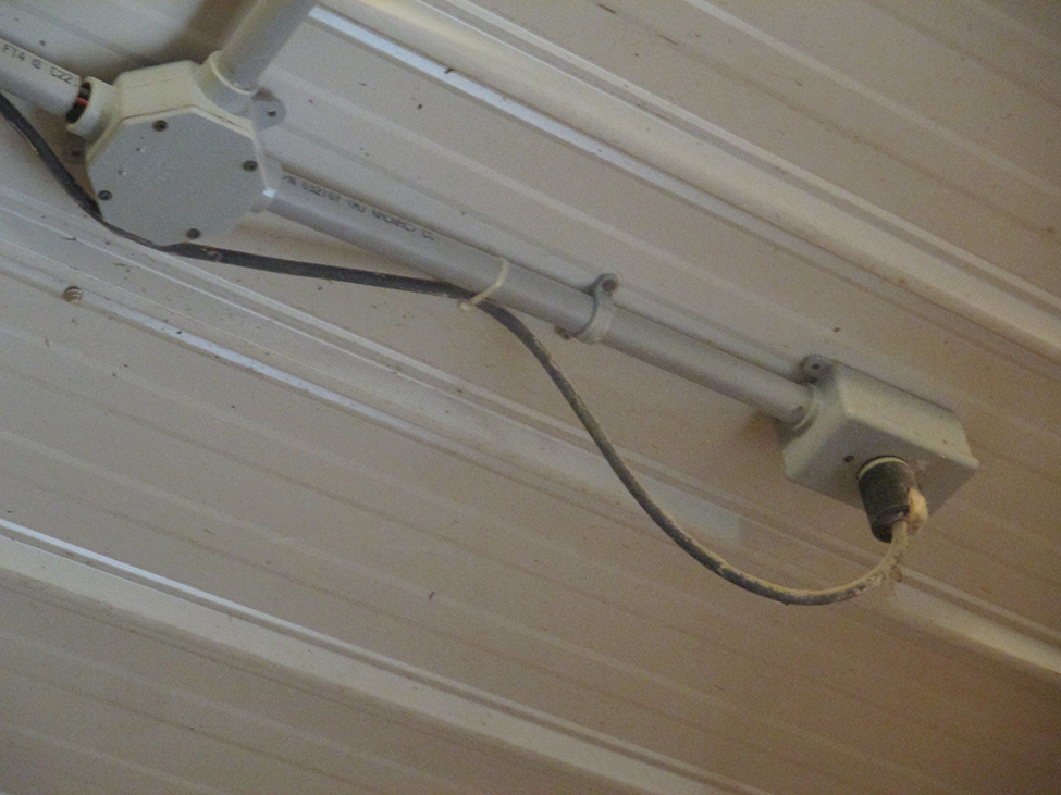 An extension cord plugged into a ceiling outlet.  Dust has accumulated on the ceiling and cord and a zip-tie is used to keep the cord in place on the electrical conduit.