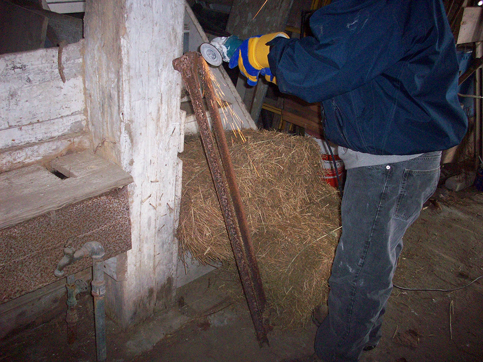 A person grinding a piece of metal inside an older livestock barn directly beside a straw bale.  Sparks are falling near the straw.