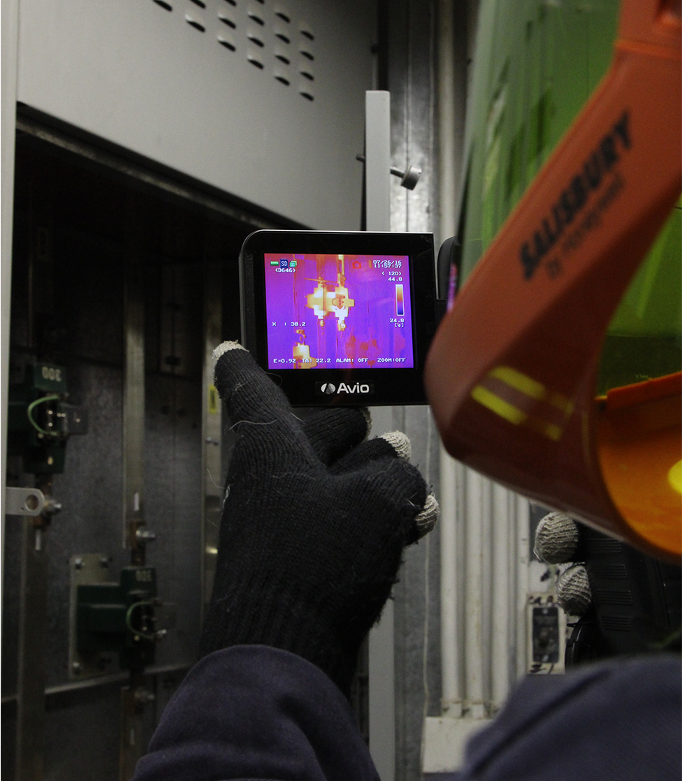 A person pointing a thermal camera at an electrical cabinet. The image on the thermal camera indicates components in the electrical cabinet are warmer (yellow) than the ambient temperature (purple).