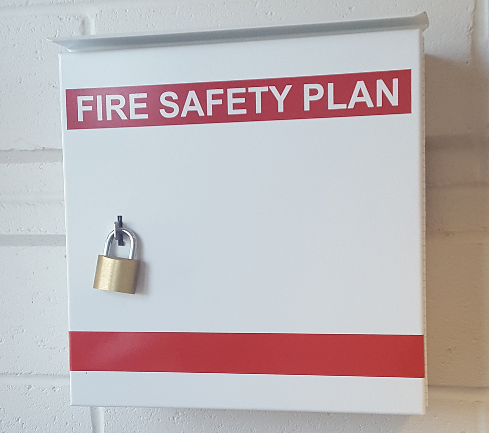 A locked, white metal box attached to a white wall with a red label that says fire safety plan.