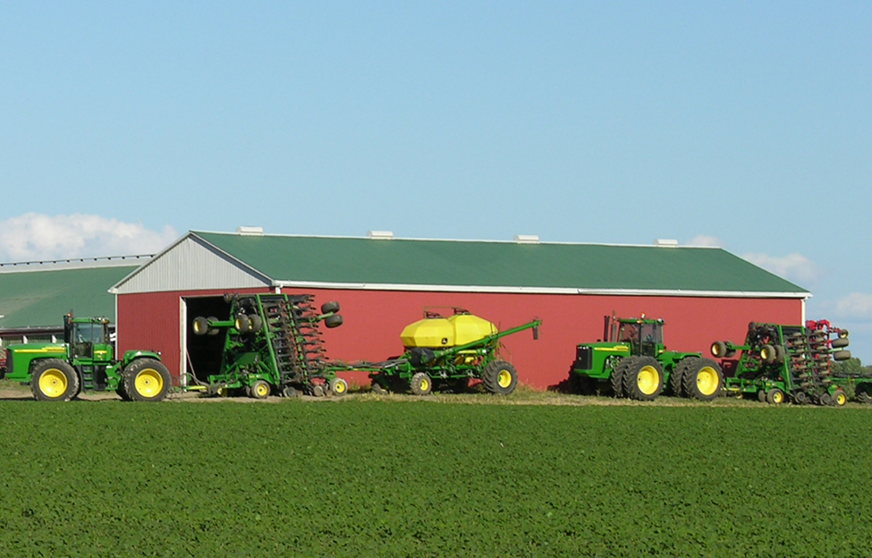 Large pieces of field equipment including five tractors, a sprayer, a wagon and two cultivators parked outside a large red equipment storage building. A moderate sized livestock barn can be seen in the background. Three large silver grain storages stand off to the left of the photo.