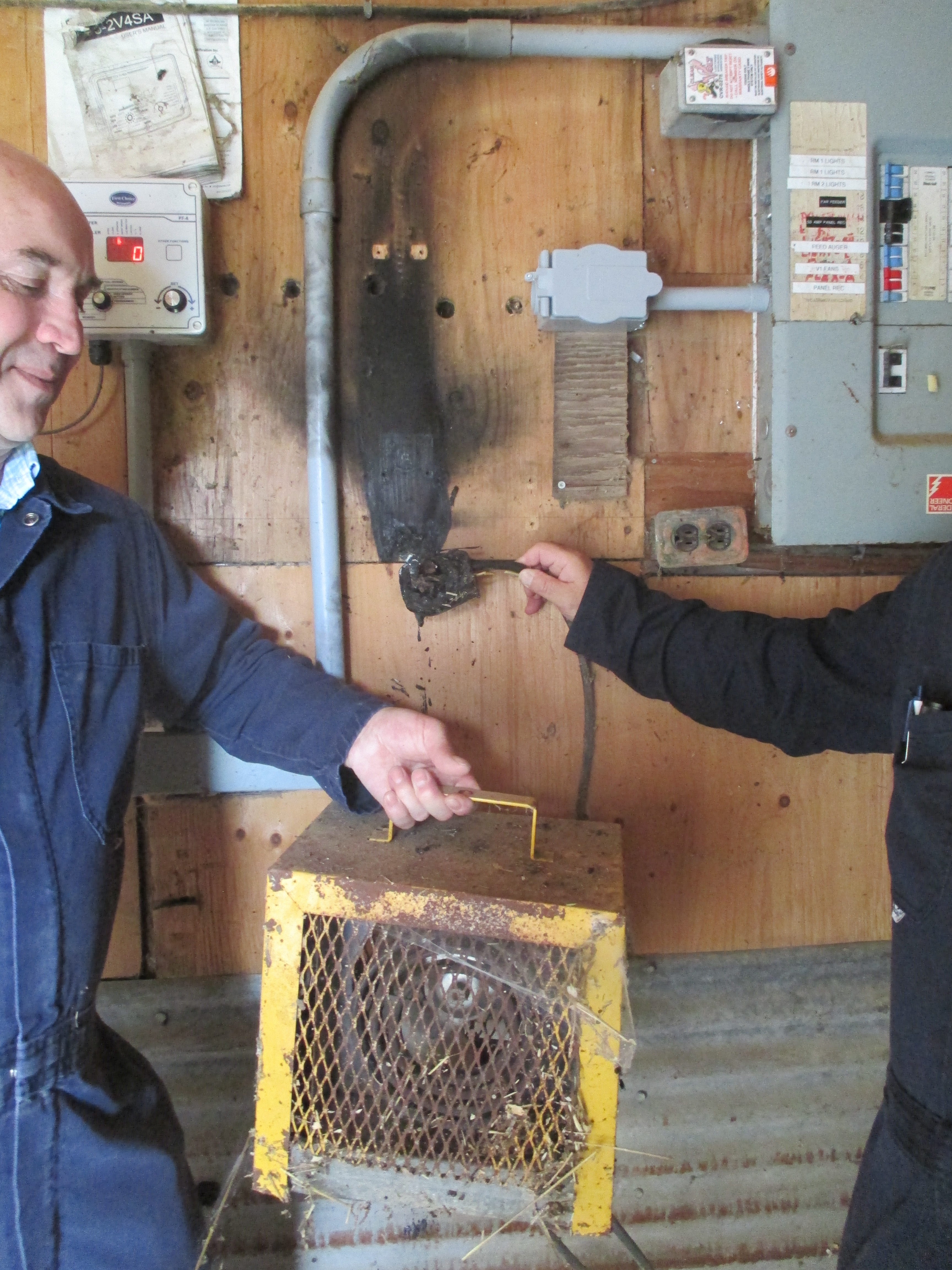 This is a photograph of box heater replaced from a barn.  The yellow box heater is rusted and there is straw visible inside the heater housing.  Electrical power was supplied to the heater through a cable and plug.  The plug is being held up against the barn wall where it used to be installed.  There is black burn marks on the plywood barn wall.  The plug itself is heavily damaged.  Its plastic housing is partially melted due to a fire that started within the plug and receptacle.