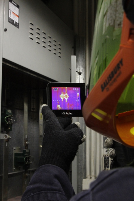 This is a photograph of an operator conducting an infrared thermal scan of an electrical panel inside a barn. The display screen of the infrared thermal scanner is visible in the photograph. The scanner displays its field of view in colors ranging from blue to white. The display color is related to temperature with blue being cool and white being hot. In this particular scan, most of the scanner display is pink in color. There are two electrical components in the center of the display which are white in color, indicating high localized heat.