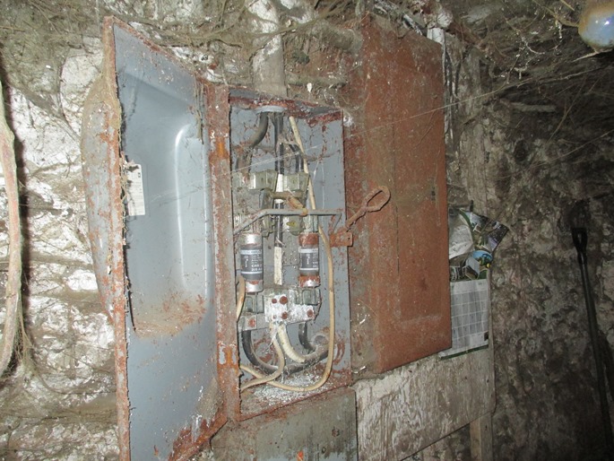 Panneau de sectionneur principal installé dans un bâtiment de ferme. Le sectionneur était sous tension lorsque la photographie a été prise. On peut voir une grande quantité de toiles d’araignées sur le mur autour du panneau. L’extérieur du panneau est rouillé, et sa porte ouverte laisse voir des fusibles et des composants fortement corrodés.