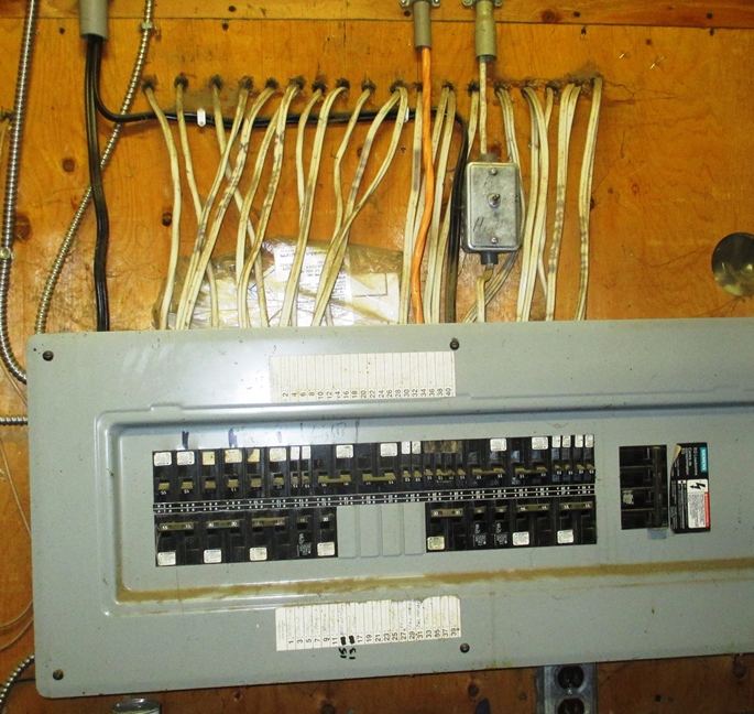 This is a photograph of a circuit breaker panel installed in a barn. The panel is of a style typically used for residential applications. The fuse panel access door has been removed and all the circuit breakers are exposed to the barn atmosphere. A large number of electrical cables exit the upper side of the circuit breaker panel. Most of these cables have been routed into holes drilled into the wall on which the panel in installed. Plastic conduit has not been used to protect the recessed wiring.