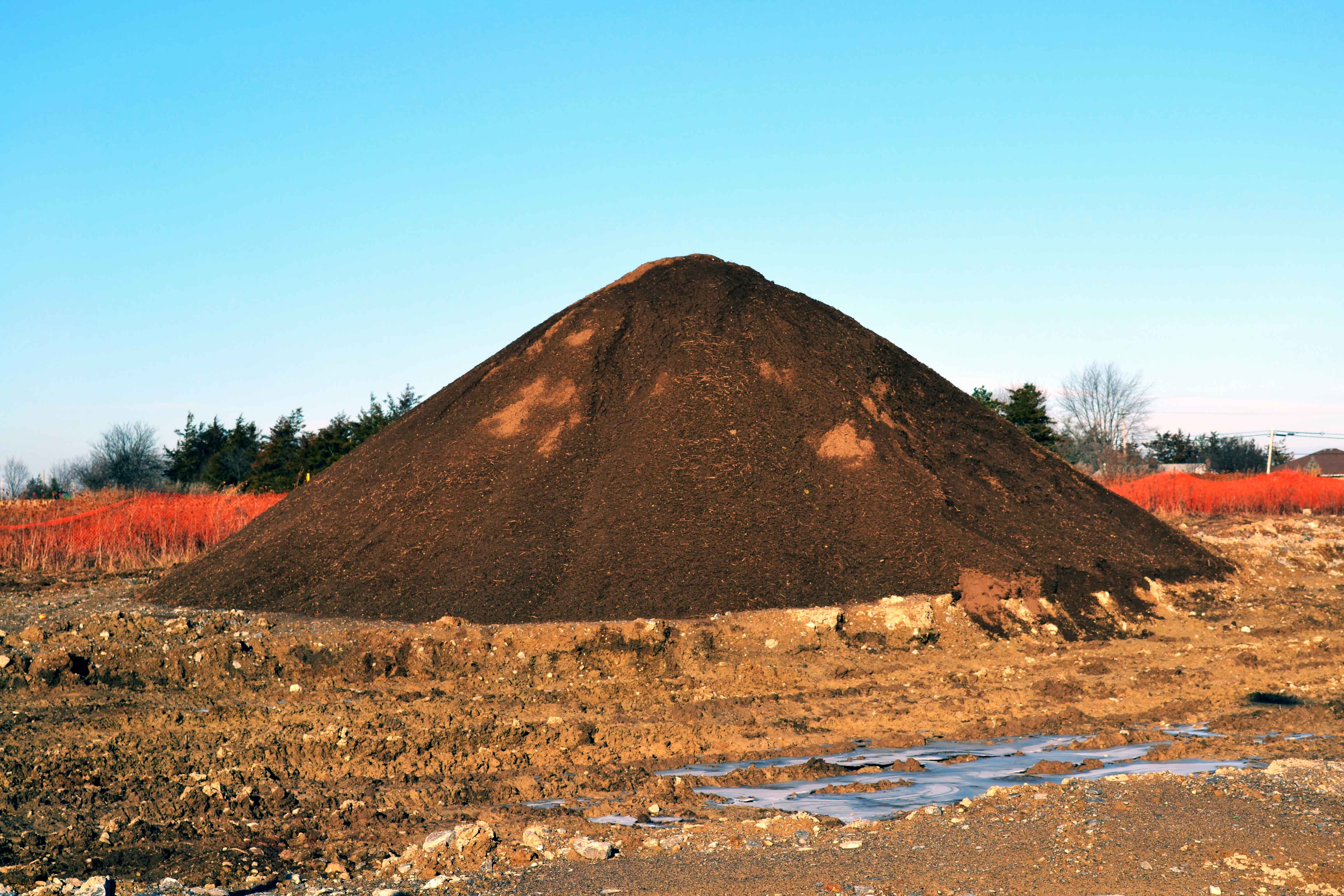 Large stockpile of brown top soil at the edge of a lot where the soil has been removed for lot development