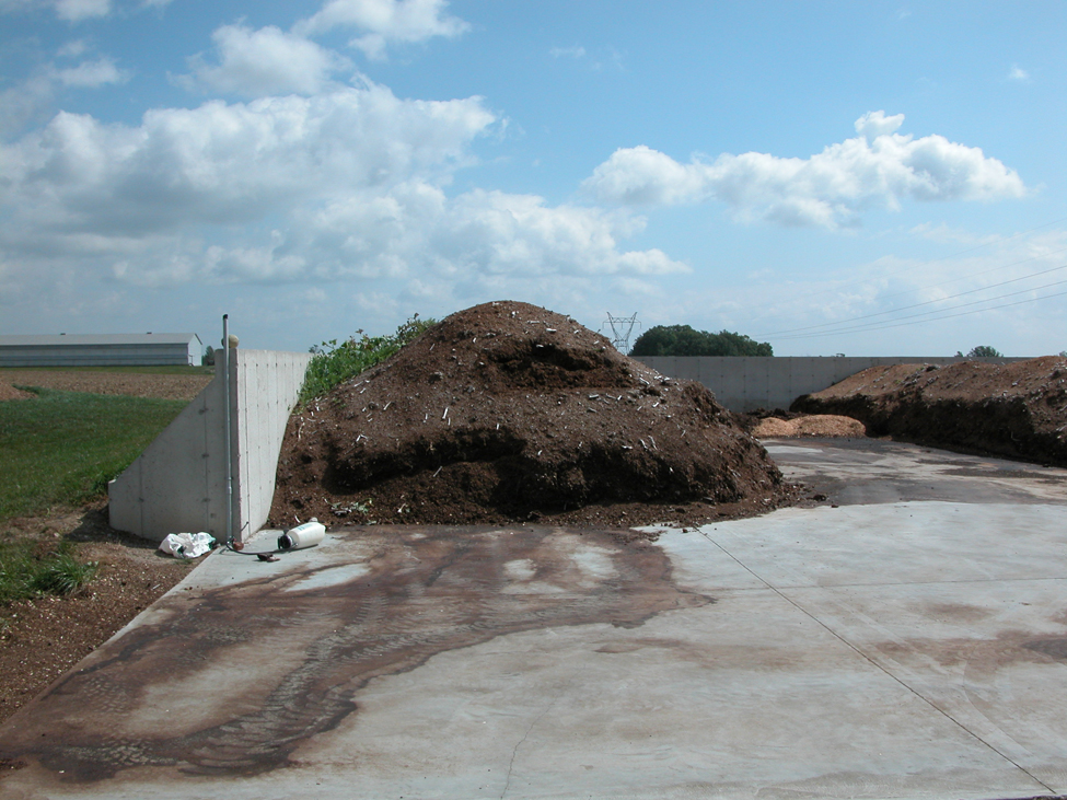 Manure piled against a concrete wall and sitting on a concrete pad. A dark brown stain shows runoff being generated from the solid manure and moving across the concrete pad.