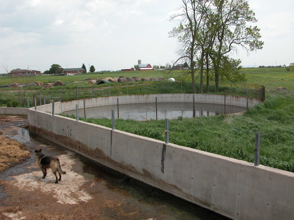 A concrete, outdoor solid manure storage yard with a fenced, circular, concrete liquid manure runoff storage tank sitting off to the right of the yard