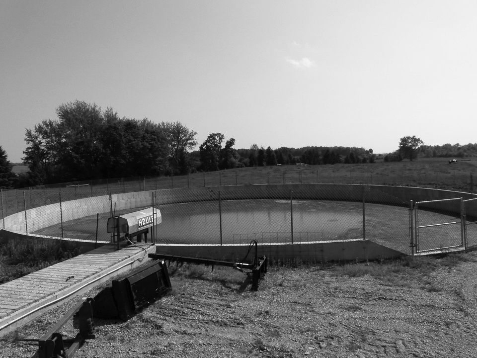An in-ground, concrete, circular manure storage tank. The tank is surrounded by a wire fence and situated next to a corn field