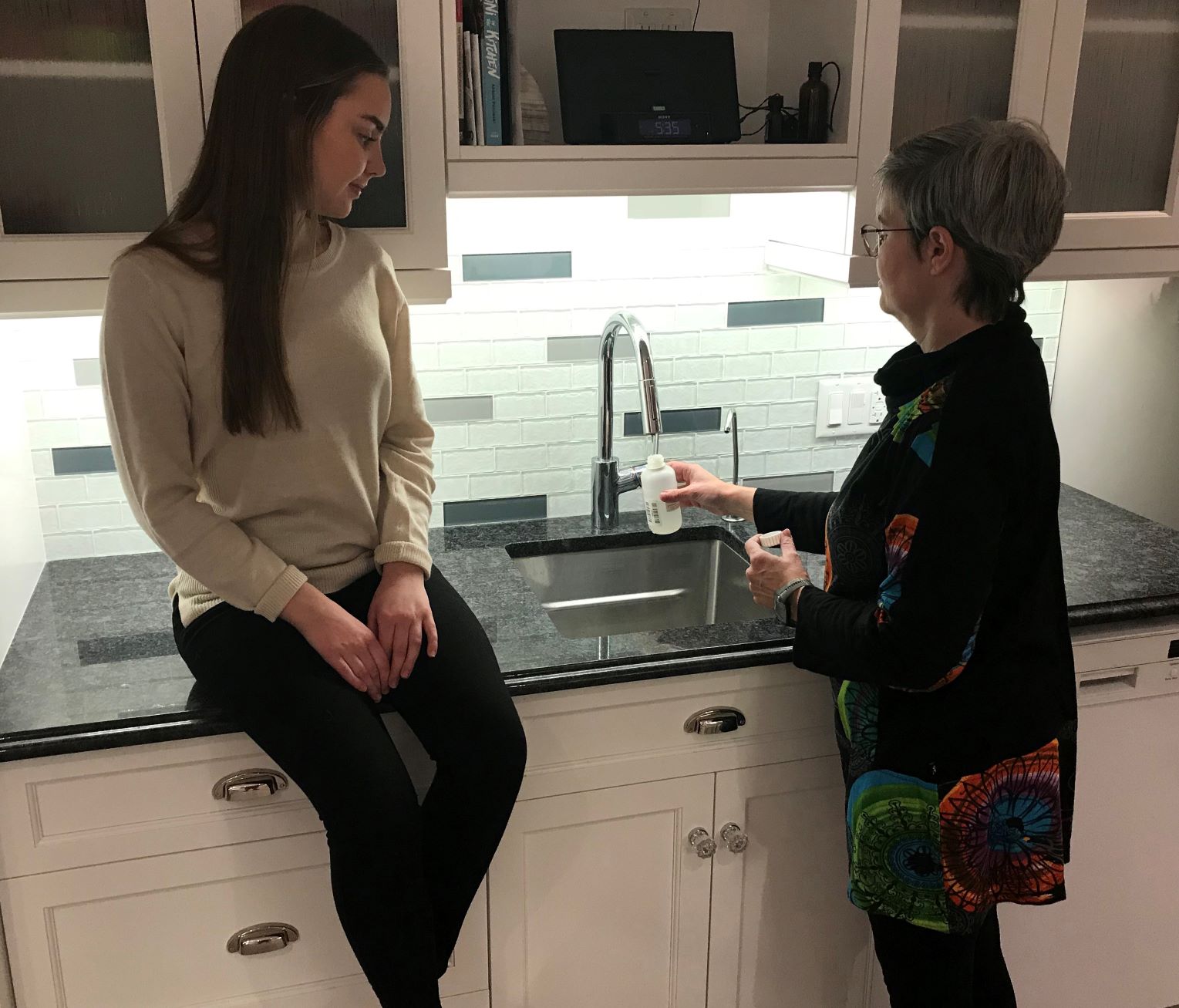 A person taking a water sample from a kitchen tap using the sample bottle provided in the water well sample kit. A second person is sitting on the kitchen countertop watching the other person take the sample. 