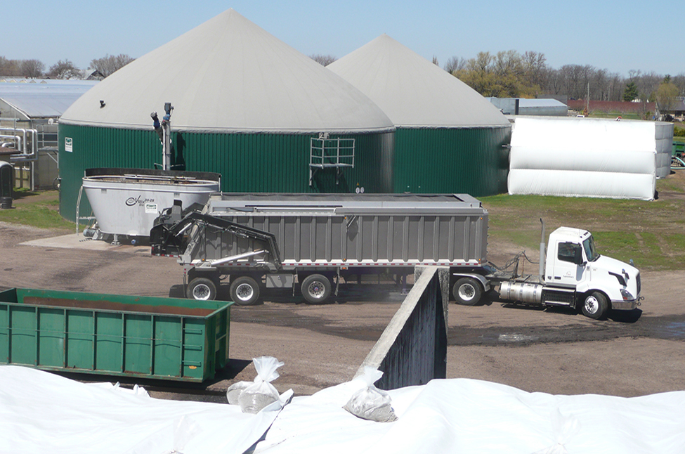large truck with a dumping trailer parked in front of a double domed anaerobic digestion system at a greenhouse facilty