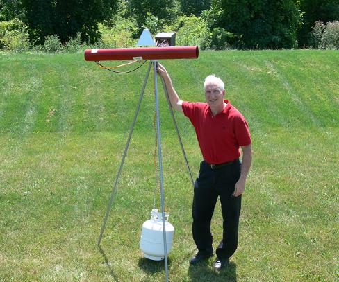 Photo montrant l'auteur debout à côté d'une bombonne de gaz alimentant un canon effaroucheur posé sur un trépied. Le canon est un cylindre rouge qui arrive à environ 30 cm au-dessus de la tête de l'auteur.