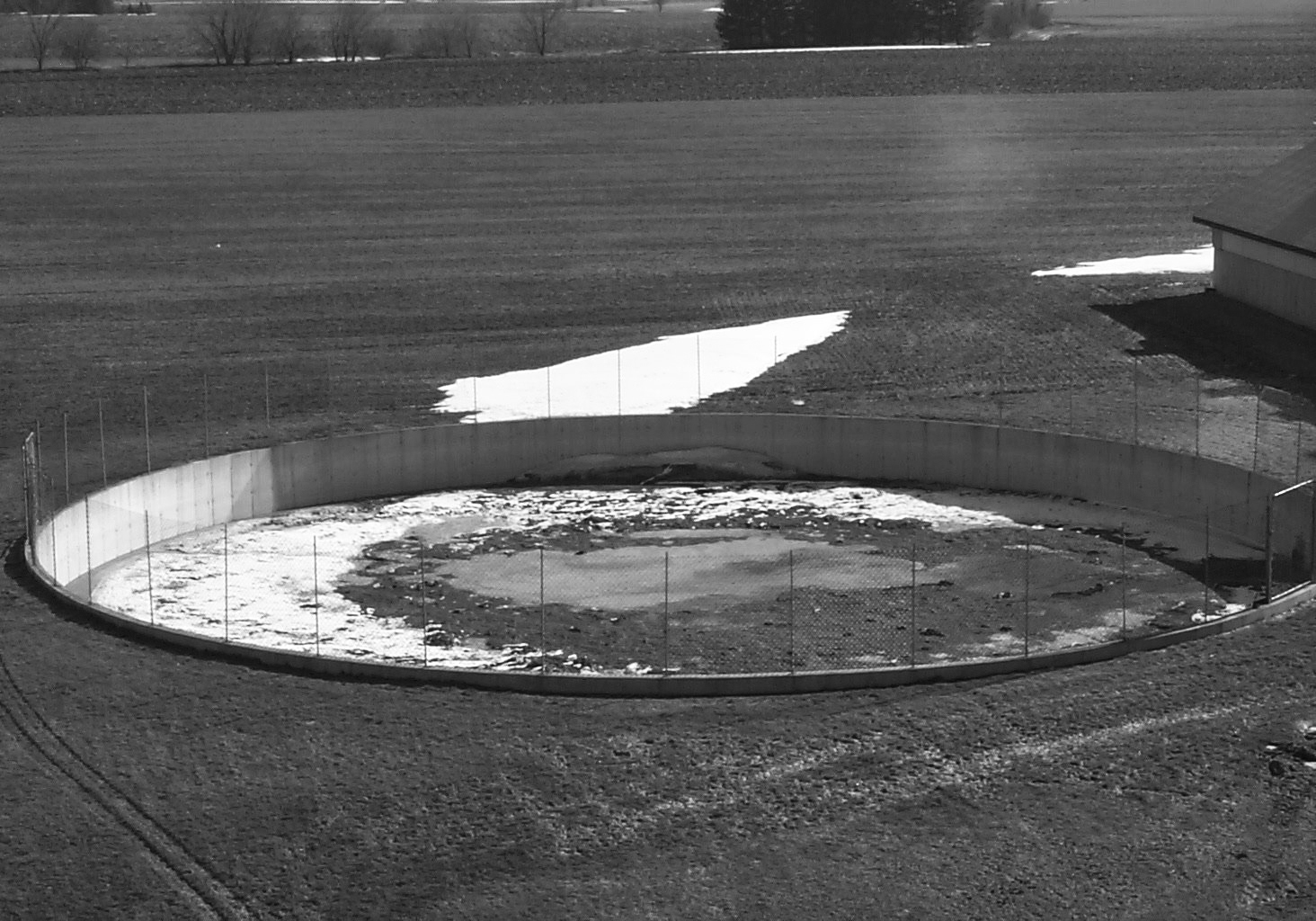 An open, circular, liquid manure tank in a field. The tank is surrounded by steel fencing.