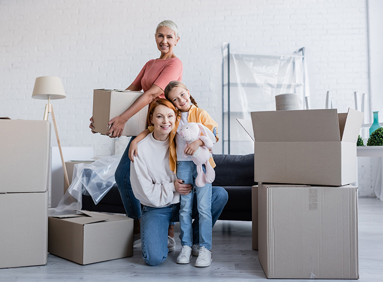 Photo of a family inside a home with moving boxes around them. One woman is lifting a box while another woman is holding their daughter.