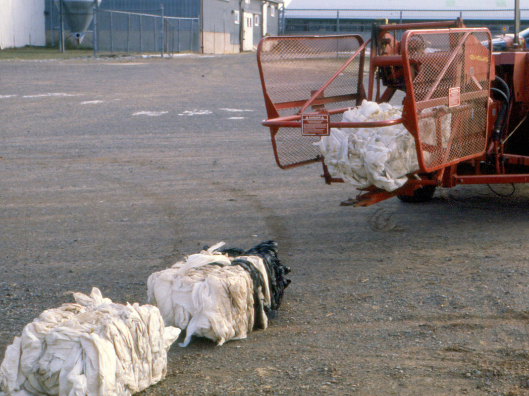 A hay baler with a recently formed white bale of scrap plastic wrap material as it exits the baler.