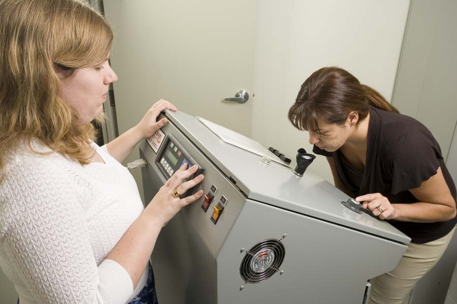 A woman standing to the left of a large grey machine where a second woman, on the right is placing her nose against the machine to assess an odour sample