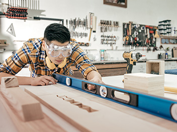 A teenager wearing safety goggles with ear protection muffs, using a leveller on a piece of wood in a workshop