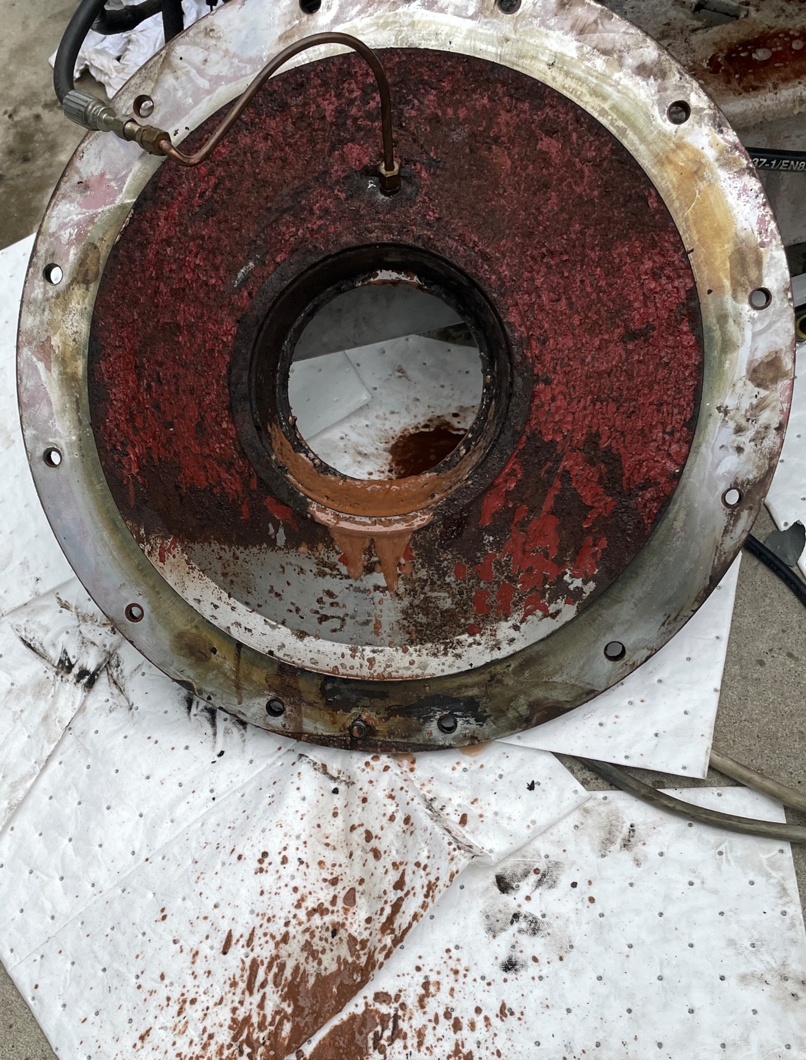 the flange that houses the roller bearing that supports the main output is removed. Pressure was connected to the lubrication system to clear the blockage in the obstructed port. Contaminated fluid is observed discharging from the lubrication system once the obstructed port is cleared