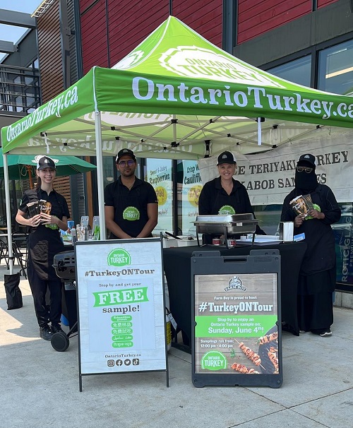 Turkey Farmers of Ontario sampling program booth that is handing out free samples local Ontario Turkey products outside of grocery stores. There are 4 people under a green tent that has OntarioTurkey.ca on the top of the tent. There are two sandwich board signs in front that say “TurkeyONTour” free sample and “TurkeyONTour” at Farm Boy. This photo was provided by Turkey Farmers of Ontario.