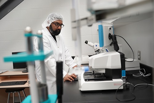 This photo shows a man wearing a hair net and white lab coat. He is working at a computer that has a microscope next to it. There is another computer desk in the background and blurred equipment in the left foreground. This photo was provided by Conestoga Food Research and Innovation Lab at Conestoga College.