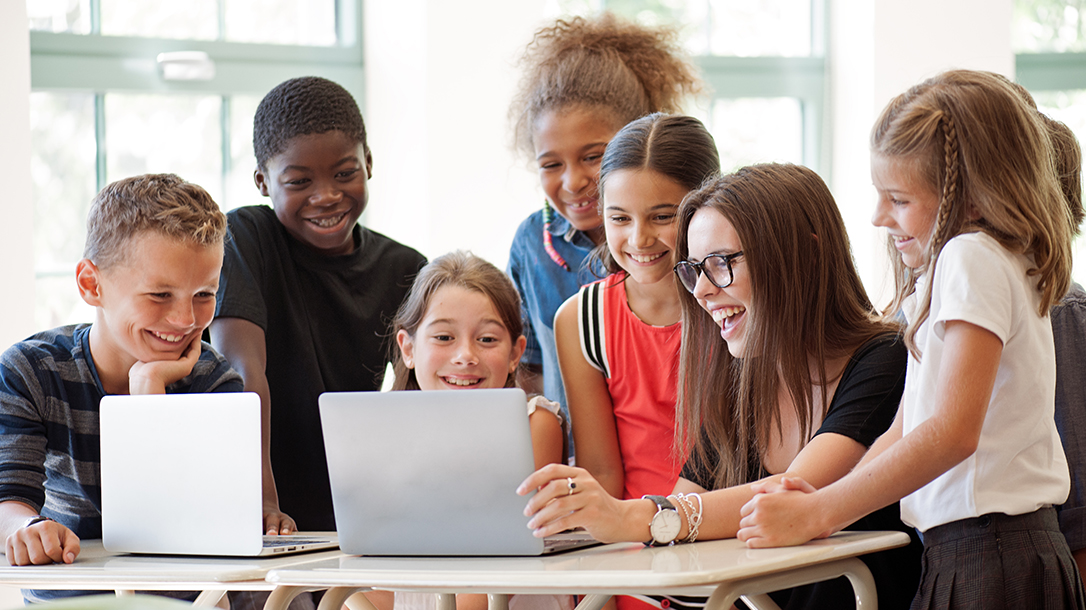 Group of students with a teacher gathered around a table with 2 notebooks. They are collaborating in a classroom setting on a focused activity