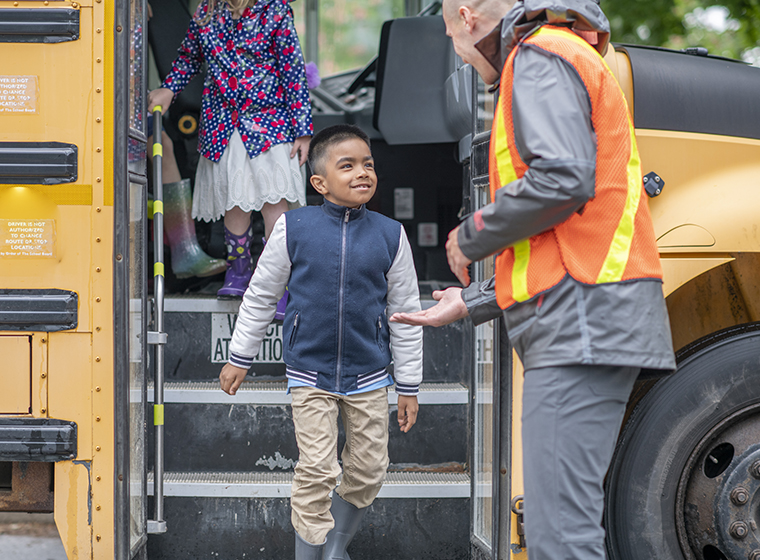 Elementary school students getting off a school bus and being greeted by school staff