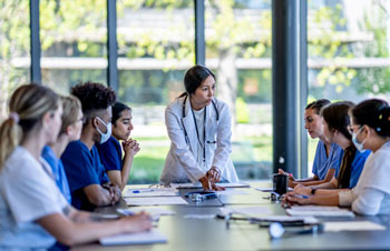 Students sitting at a conference table wearing medical scrubs, being spoken to by a supervisor wearing a lab coat and a stethoscope.