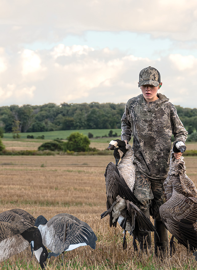 Front cover of the 2025 of the Hunting Regulations Summary with a boy walking in a field carrying two geese and geese decoys in front of him.