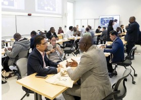 Photo d’une grande salle où plusieurs groupes de personnes sont assis autour de tables, discutant en binômes ou petits groupes ; on aperçoit au fond un écran de présentation affichant du contenu.