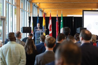 Photo prise lors de la mission commerciale de l’OIF coparrainée par l’Ontario : Dominic Mailloux, président de la Fédération des gens d’affaires de l’Ontario, debout à un pupitre face à l’audience, avec derrière lui plusieurs drapeaux de provinces et pays, et un écran de projection partiellement visible à droite.