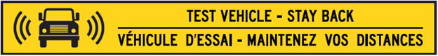 A yellow sign with an image of an automated commercial vehicle and text in black reading "Test vehicle - stay back" in English and "Véhicule d'essai - maintenez vos distances" in French