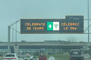 Electronic roadside message board overlooking the roadway, displaying a message celebrating the 50th anniversary of the Franco‑Ontarian flag. The message reads: “Celebrate 50 years / Célébrez le 50ᵉ.”