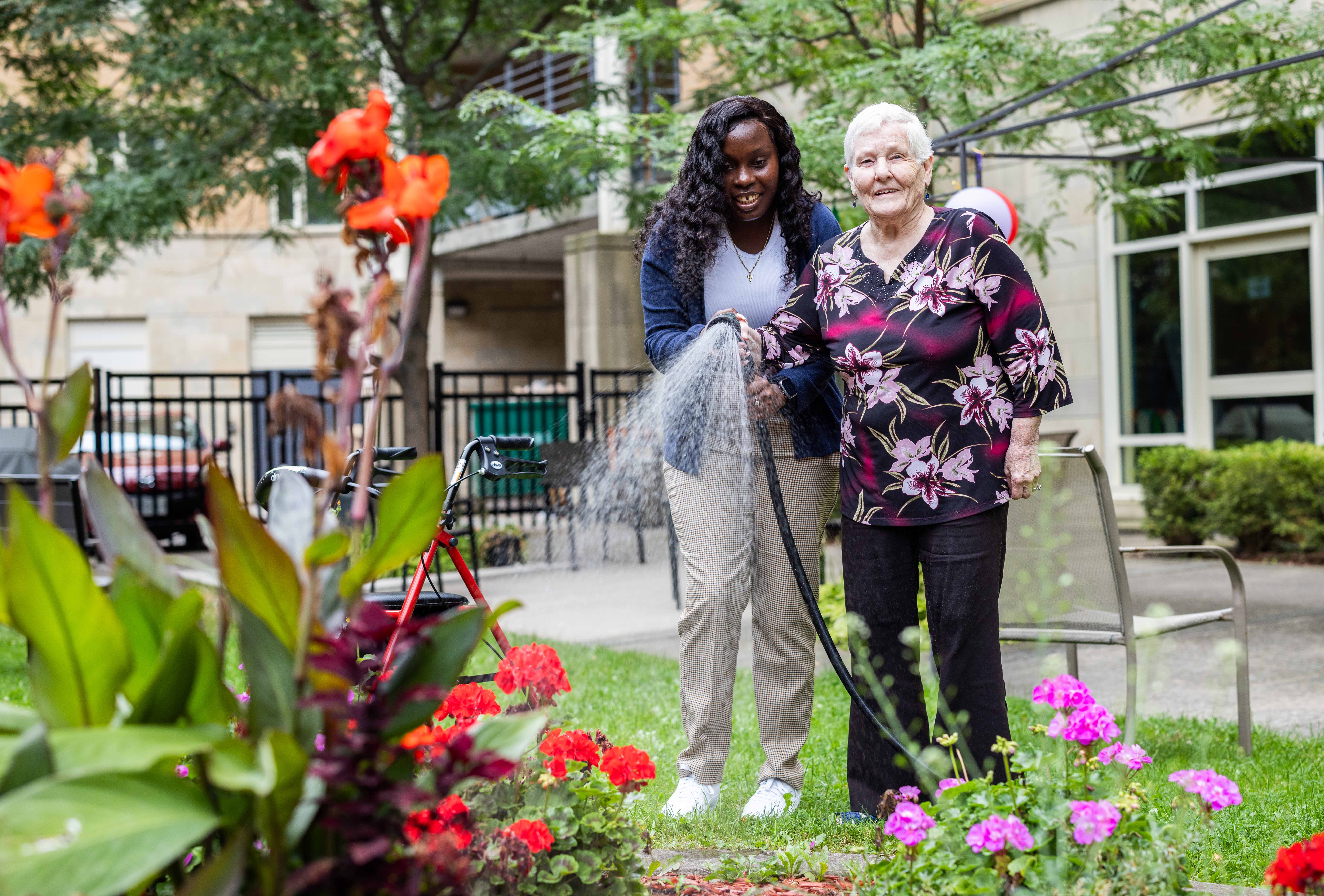Two people water colorful flowers together in a lush garden outside a residential building.