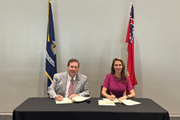 Photographie de la ministre des Affaires francophones de l’Ontario, l’honorable Caroline Mulroney, vêtue d’une robe rose, et du lieutenant-gouverneur de la Louisiane, Billy Nungesser, portant un costume gris, assis côte à côte à une table tout en tenant une déclaration conjointe signée, avec les drapeaux de l’Ontario et de la Louisiane en arrière-plan.
