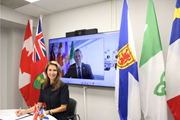 Photographie de la ministre Caroline Mulroney, vêtue d’une robe noire, assise devant une table blanche alors qu’elle signe une déclaration conjointe. Derrière elle, le ministre des Affaires acadiennes et de la Francophonie du gouvernement de la Nouvelle‑Écosse, Colton LeBlanc, apparaît à l’écran. À côté de l’écran se trouvent les drapeaux du Canada, de l’Ontario, des Franco‑Ontariens, de la Nouvelle‑Écosse et de l’Acadie.