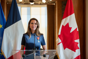 L’Honorable Caroline Mulroney, ministre des Affaires francophones,  prenant la parole à la tribune, avec les drapeaux français et canadiens en arrière‑plan, lors de sa décoration de la Légion d’honneur.