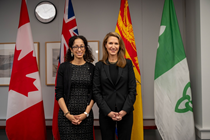 Photographie de la ministre des Affaires francophones de l’Ontario, l’honorable Caroline Mulroney, et de la sous‑ministre adjointe Maria Lamani, debout côte à côte, toutes deux vêtues de noir, avec les drapeaux du Canada, de l’Ontario, des Franco‑Ontariens, de l’Acadie et du Nouveau‑Brunswick en arrière‑plan.
