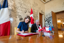 Photographie de la ministre des Affaires francophones de l’Ontario, l’honorable Caroline Mulroney, et de l’ambassadeur de France au Canada, Michel Miraillet, assis côte à côte à une table en bois et signant tous deux une déclaration conjointe. De petits drapeaux sont disposés sur la table, tandis que les drapeaux du Canada et de la France se trouvent en arrière‑plan.