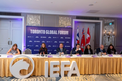 Photographie de la ministre des Affaires francophones de l’Ontario, l’honorable Caroline Mulroney, assise à une longue table avec Jean‑Claude D’Amours, ministre du Cabinet du Nouveau‑Brunswick, lors du Forum mondial de Toronto, en compagnie d’autres représentants. Un grand panneau affichant les lettres « IEFA » est placé devant la table.