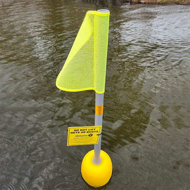 Une bouée jaune avec un poteau et un drapeau jaune flotte à la surface du lac. Une étiquette attachée au poteau informe toute personne qui la consulte que le filet a été installé par le personnel du gouvernement pour la surveillance des pêches.